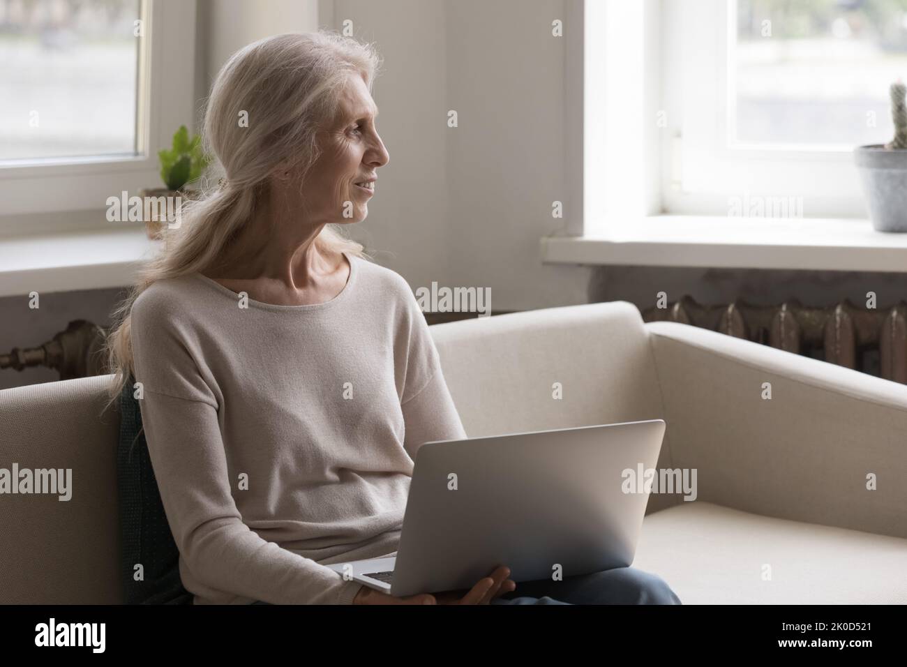 Donna anziana felice e sognante che tiene il laptop in braccio, guardando via Foto Stock