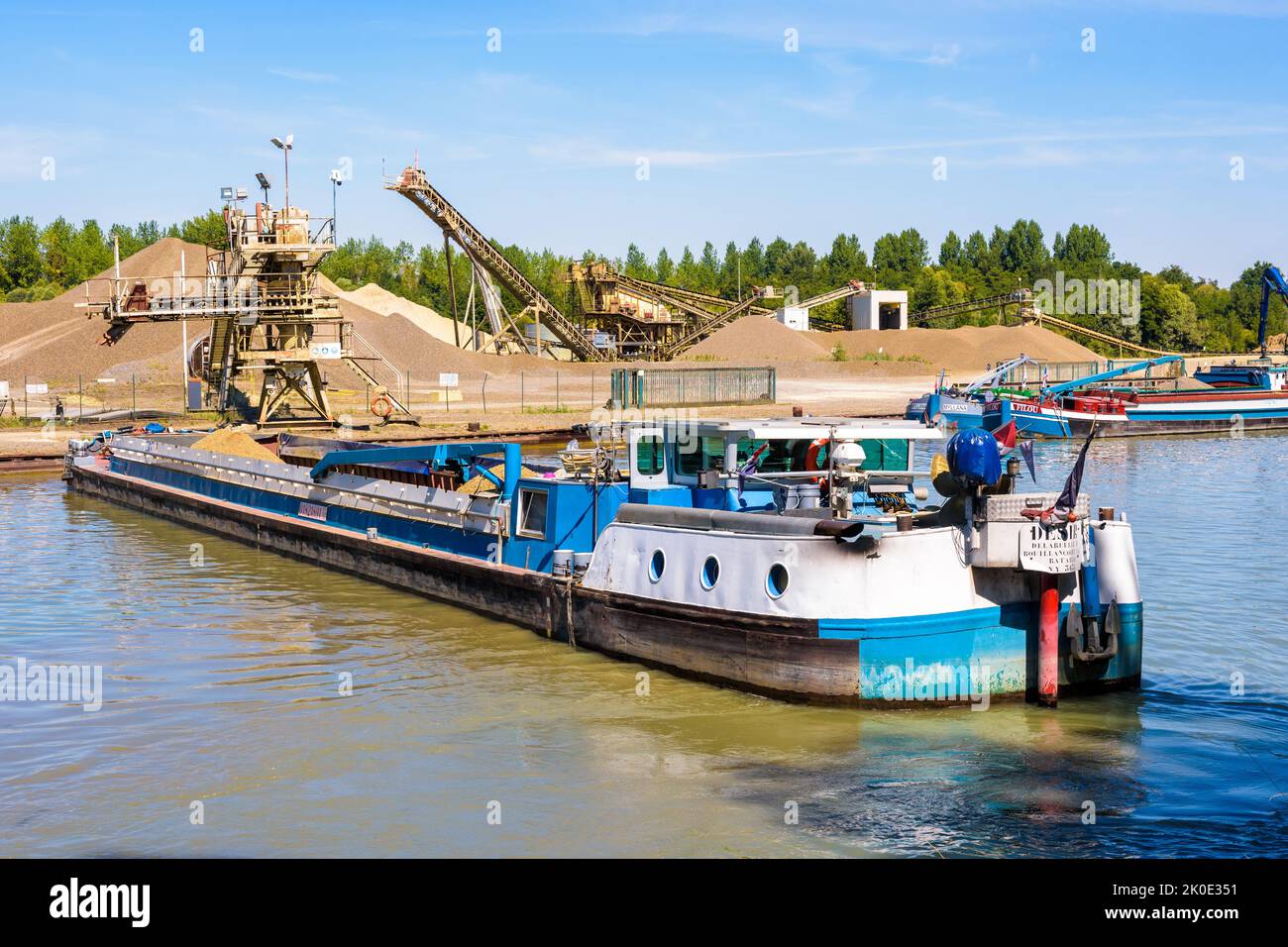 Una chiatta motorizzata carica di ghiaia si sta manovrando su un canale per ormeggiare alla cava di sabbia in una giornata di sole estate. Foto Stock