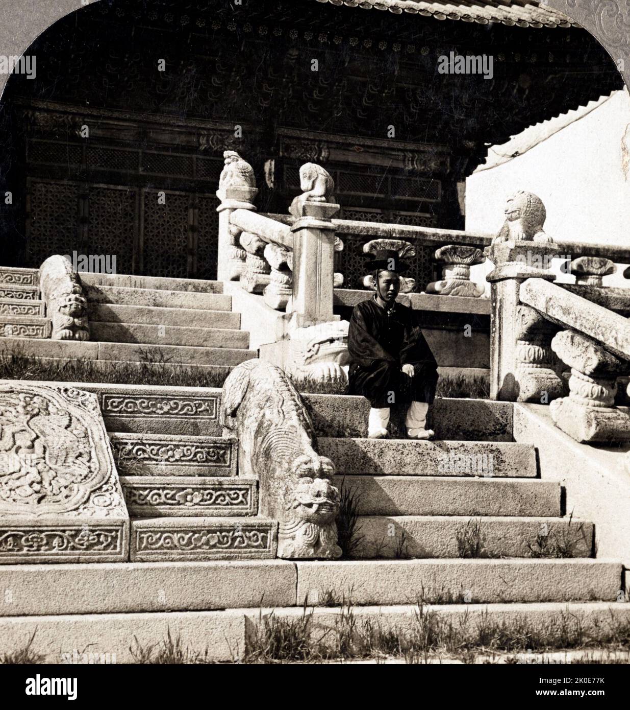 Joseon era, Corea, 1890-1900. Un servo del Palazzo reale Coreano seduto sulle scale di un edificio all'interno del Palazzo reale a Seoul. Foto Stock