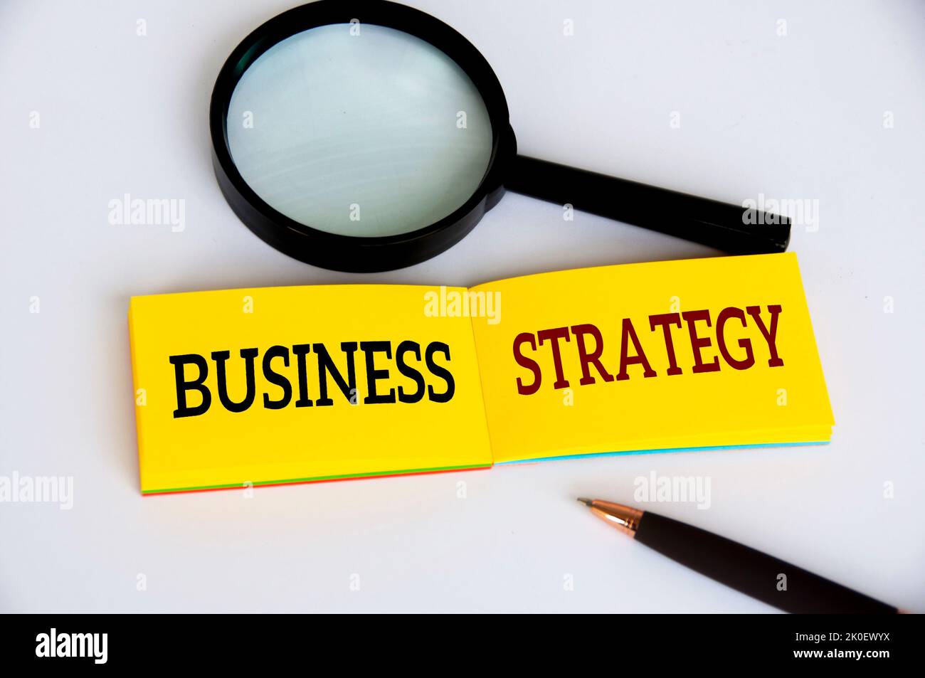 Testo di strategia aziendale su blocco note giallo con lente di ingrandimento e penna su sfondo bianco. Concetto di strategia aziendale. Foto Stock