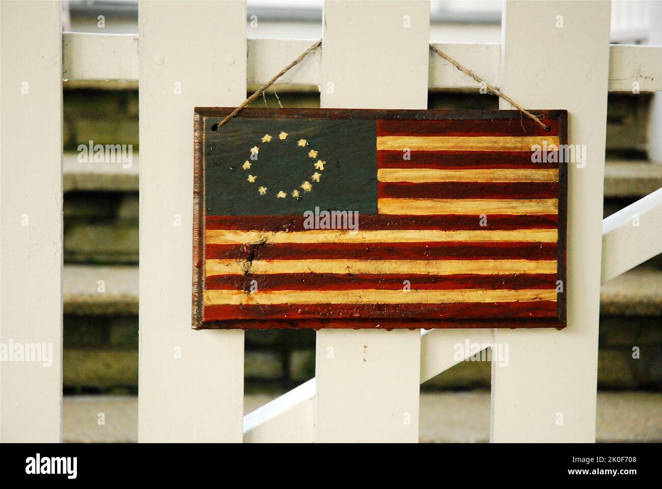 Una bandiera coloniale americana, fatta di vernice e legno, è appesa da un recinto bianco di una casa il 4 luglio Foto Stock