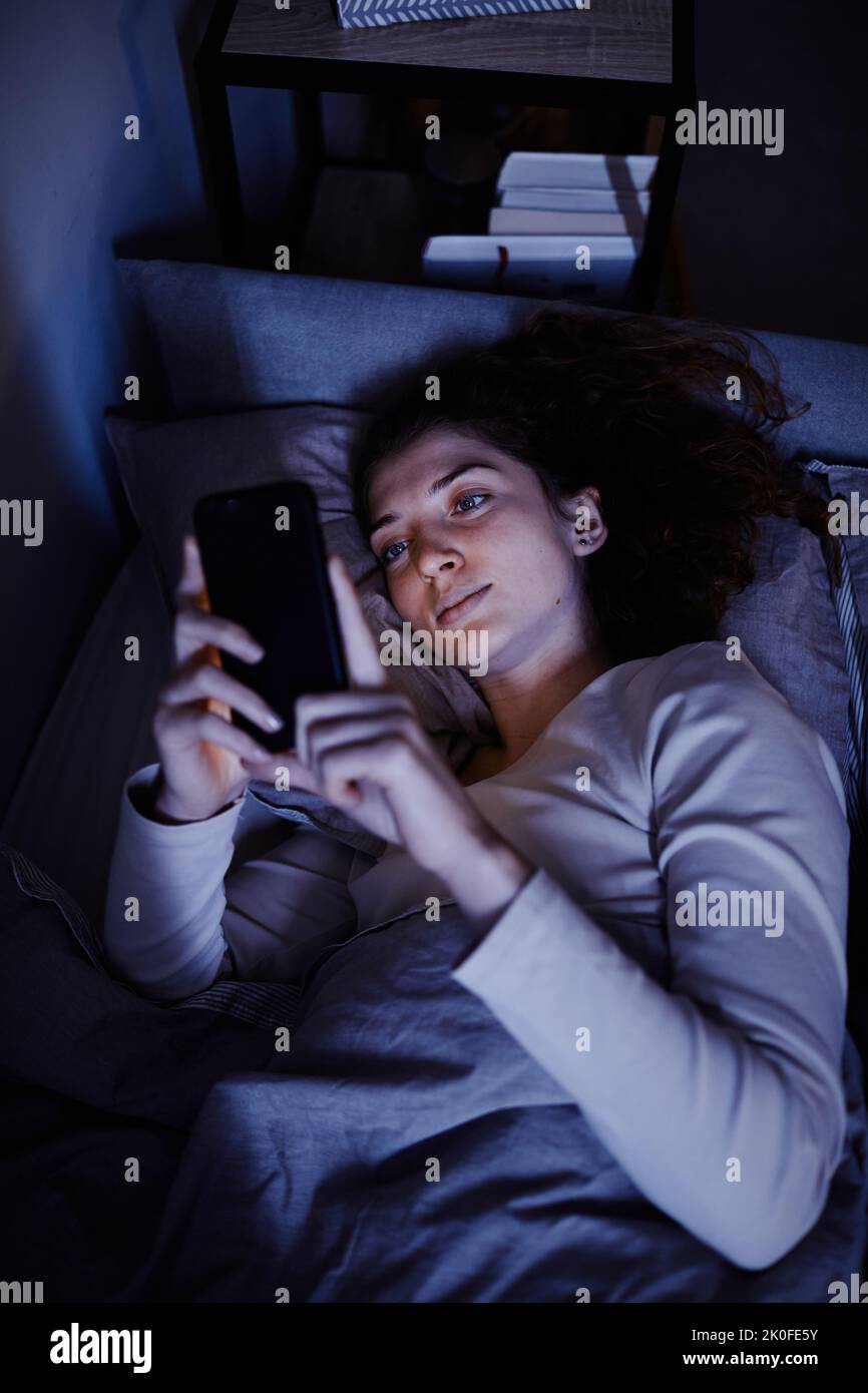 Vista ad angolo alto della giovane donna che comunica online sul telefono cellulare prima di dormire sdraiata sul letto Foto Stock
