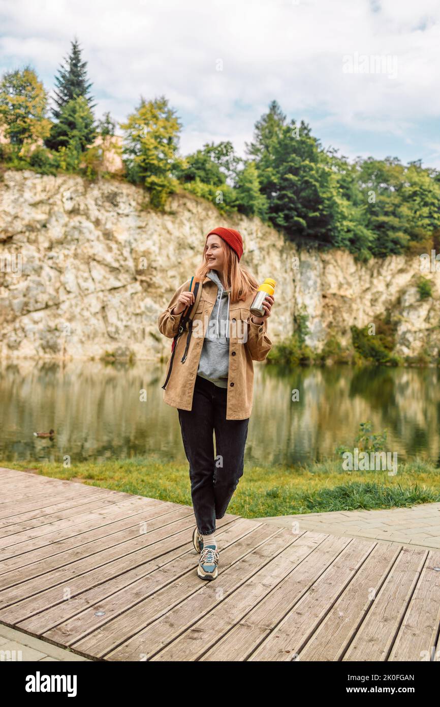 Giovane donna sorridente zaino in spalla felice con capelli biondi e beanie rosso sorride sulla macchina fotografica in cima montagne rocciose lago. Avventura, viaggio, vacanza Foto Stock