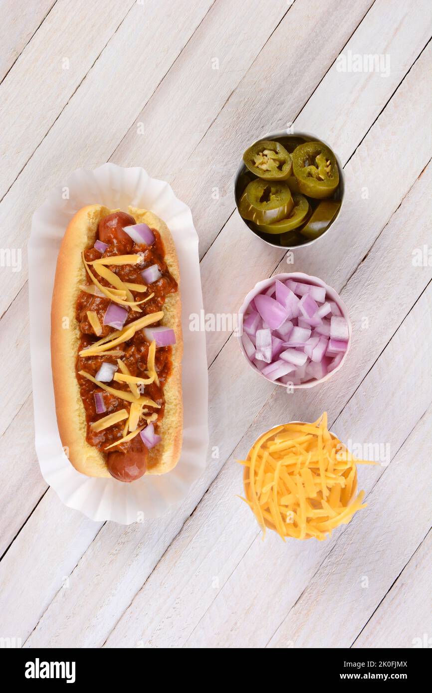 Shot ad angolo alto di un Chili Hot Dog con peperoncino, cipolle e formaggio grattugiato su un tavolo da picnic rustico in legno bianco. Tazze di cipolle tritate, peperoni e che Foto Stock