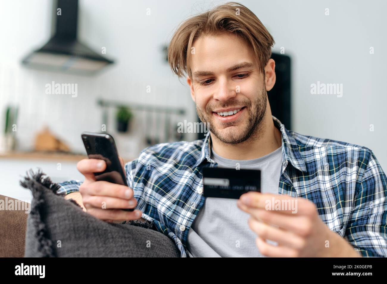 Positivo soddisfatto ragazzo caucasico sorridente, in abiti casual elegante, si siede sul divano a casa, tiene in mano il telefono cellulare e la carta di credito, paga per gli acquisti online, inserisce un numero di carta, shopping online Foto Stock