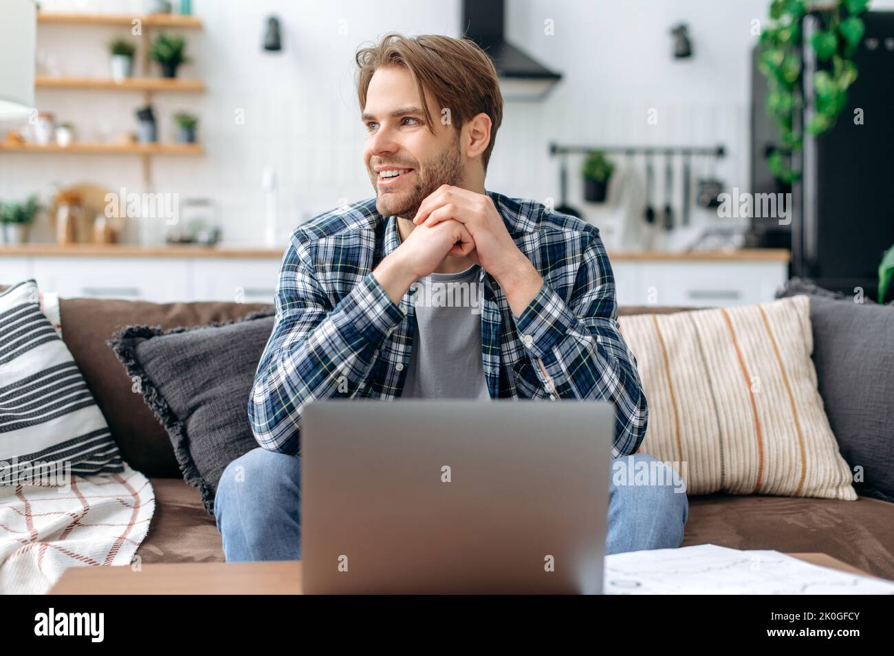 Soddisfatto bel giovane uomo caucasico in abiti casual elegante, seduto sul divano a casa in un accogliente soggiorno, utilizzando un computer portatile, lavorando o studiare online, guardando al lato, sognando, sorridendo Foto Stock