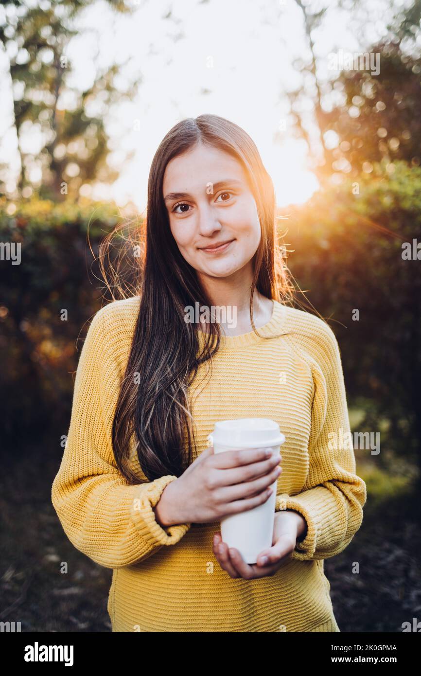 Giovane ragazza sorridente che indossa un maglione giallo e tiene una tazza di caffè usa e getta al tramonto nel parco. Atmosfera autunnale Foto Stock