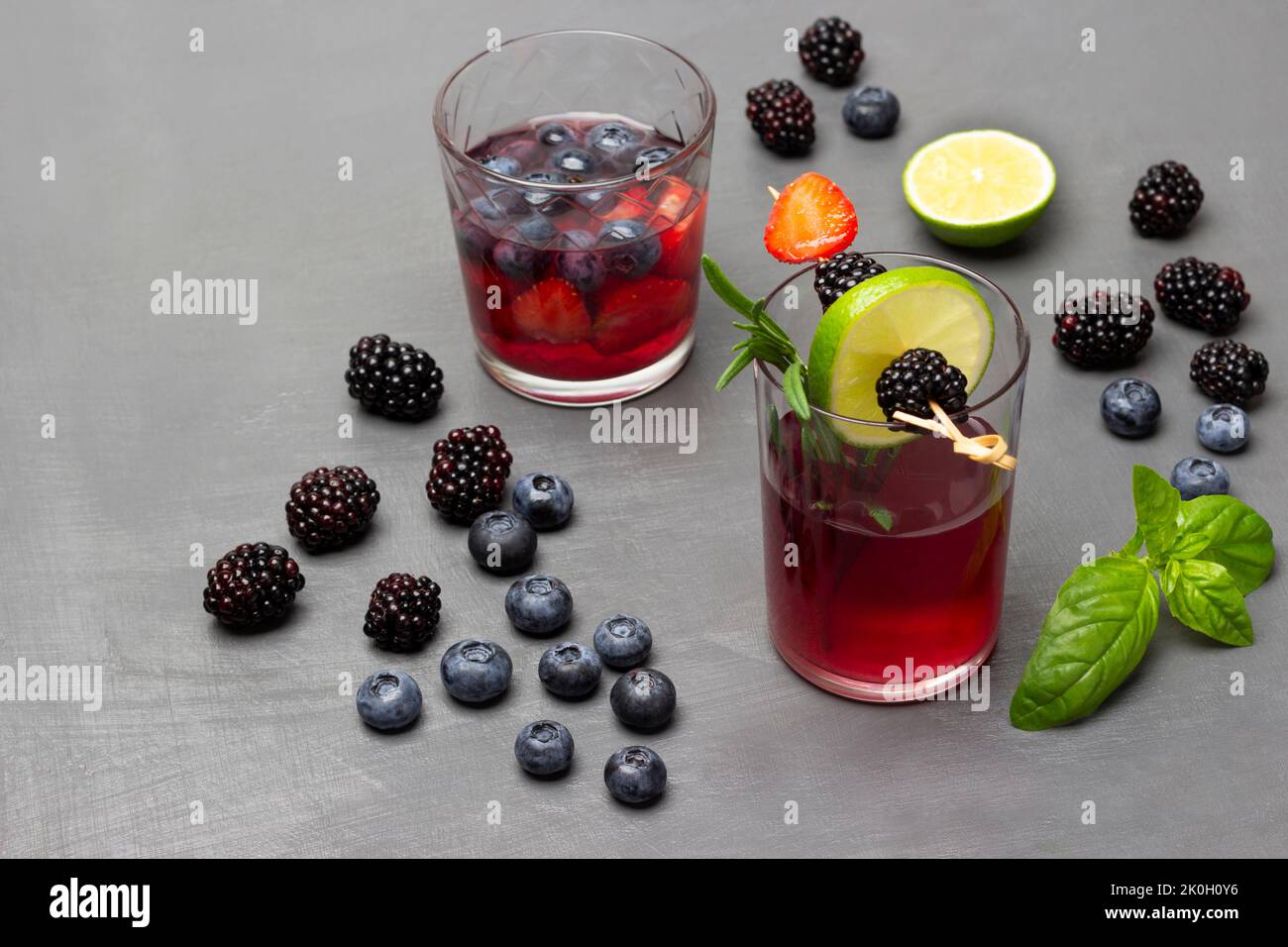 More, mirtilli, foglie di basilico verde sul tavolo. Due bicchieri di bevanda ai frutti di bosco. BlackBerry e lime cuneo su spiedino. Vista dall'alto. Sfondo grigio. Foto Stock