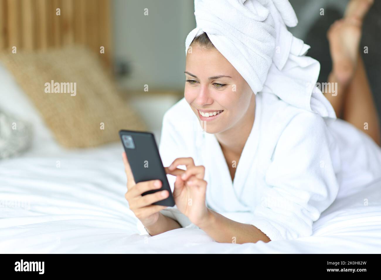 Donna felice dopo aver fatto la doccia utilizzando il telefono cellulare sdraiato sul letto a casa Foto Stock