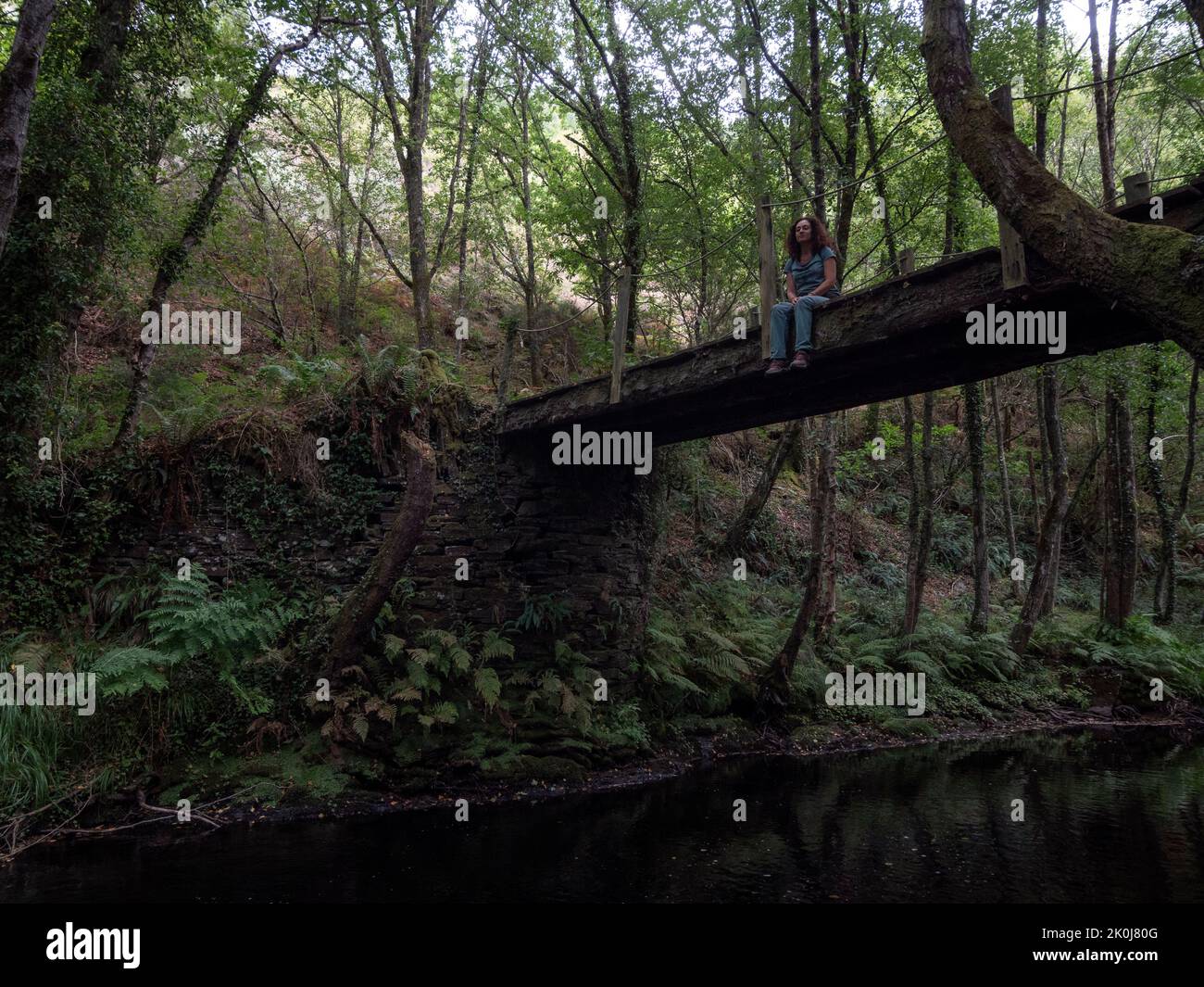 Vista orizzontale della donna brunetta seduta su un ponte di legno con fiume che scorre attraverso un querce e castagni sullo sfondo. Foto Stock