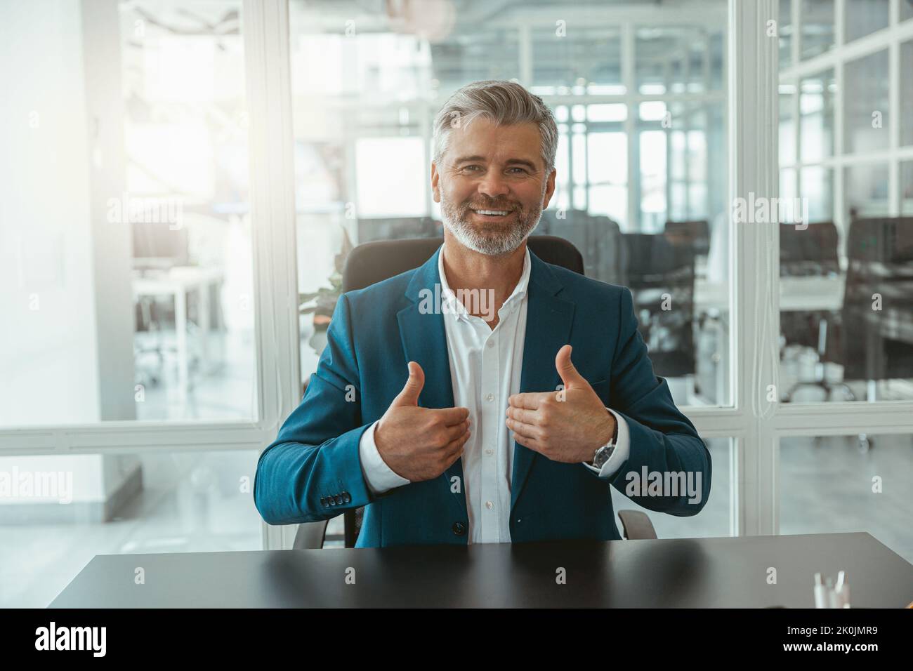 Uomo d'affari maturo sorridente che mostra le dita sollevate mentre si siede in ufficio Foto Stock