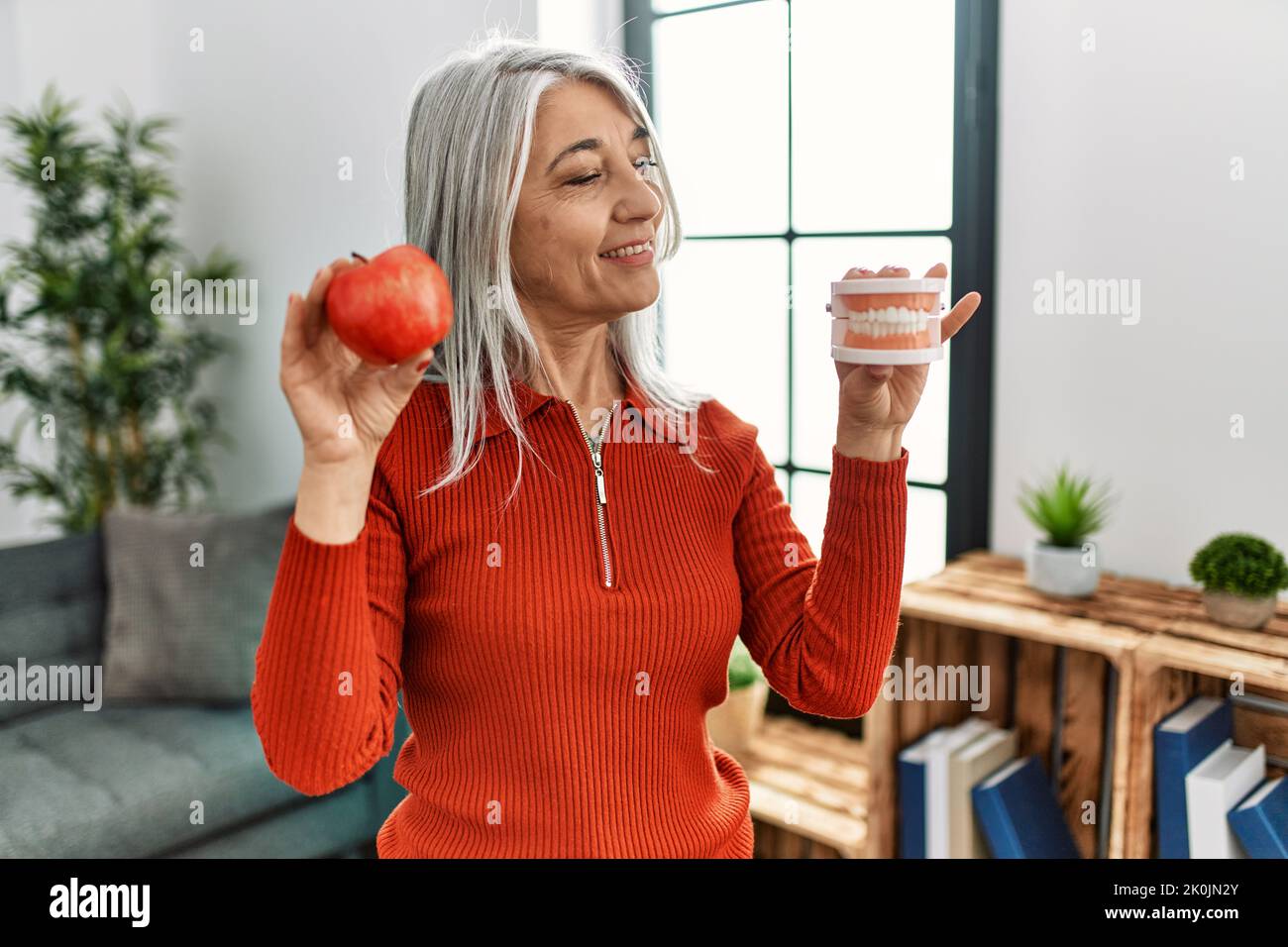 Donna di mezza età dai capelli grigi sorridente sicura che tiene la mela rossa e la dentiera a casa Foto Stock