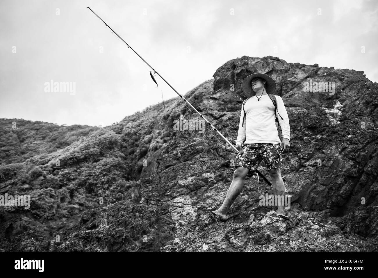Corpo pieno di uomo bianco e nero in cappello con canna da pesca con spina in piedi sulla scogliera rocciosa sulla riva, mentre la pesca in natura Foto Stock