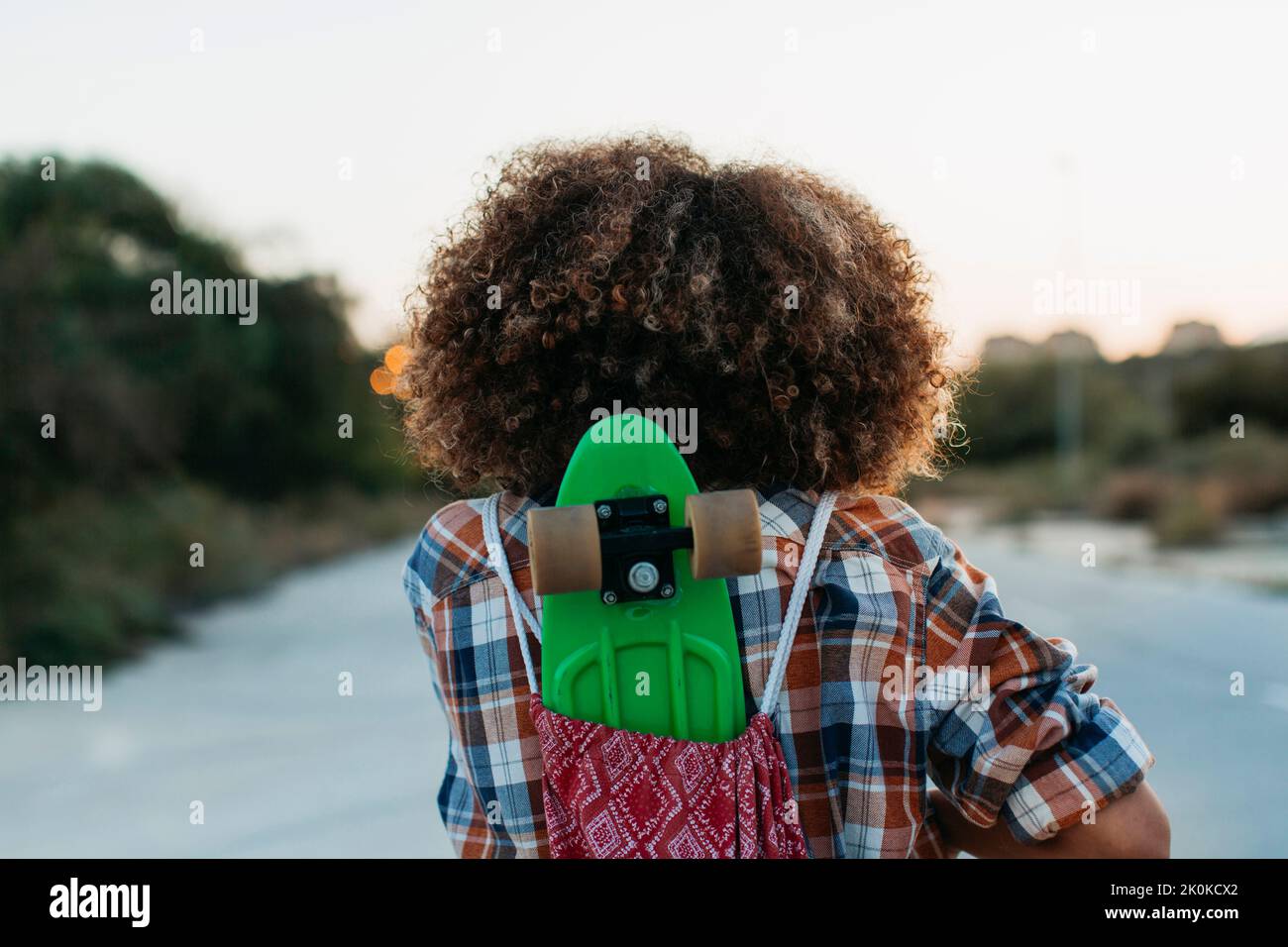 Vista posteriore del pattinatore femminile in camicia a scacchi in piedi su strada vuota con penny board verde e ammirare il tramonto Foto Stock