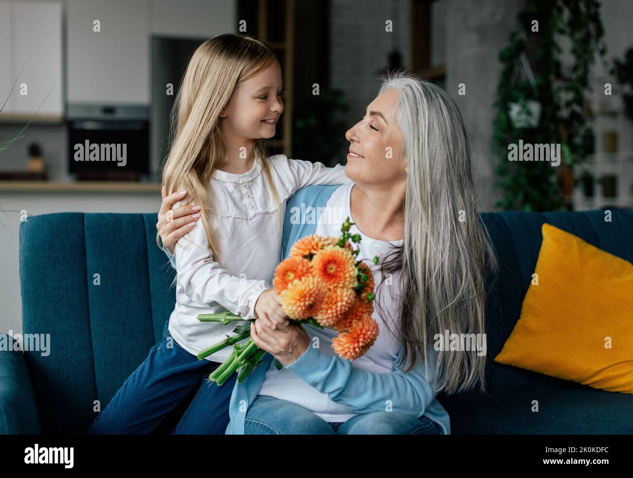 Allegra bambina abbraccia l'anziana signora europea, dà fiori e congratulazioni, divertirsi in soggiorno Foto Stock