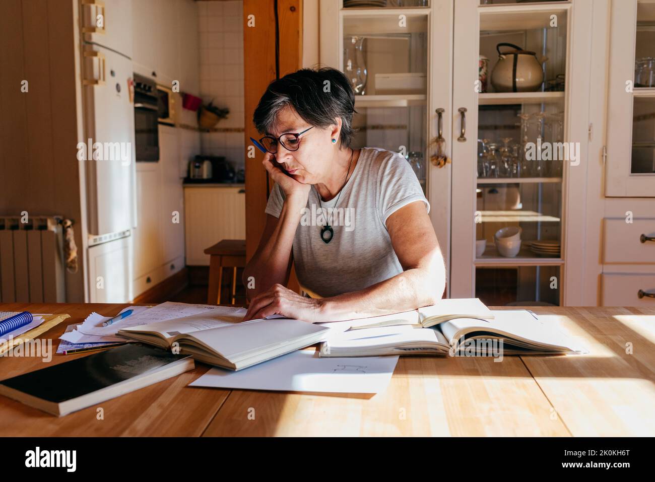 Donna di mezza età messa a fuoco in occhiali con penna in mano seduta a tavolo con un mucchio di libri e carte e studiare materiali professionali a casa Foto Stock