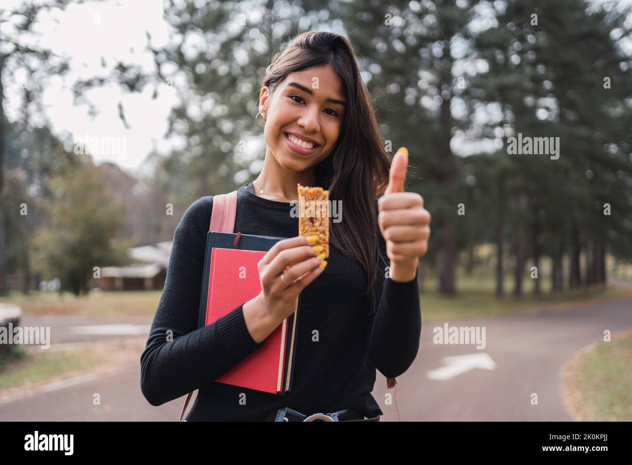 Soddisfare giovane studentessa ispanica con libri che mostrano pollice in su gesto mentre godendo sano barretta di cereali durante la pausa nel parco verde nel campus Foto Stock