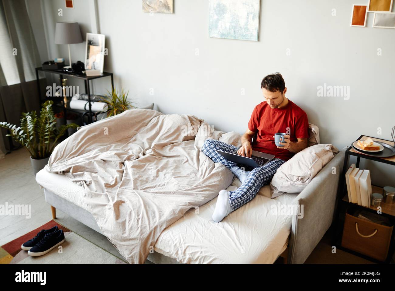 Vista ad angolo alto del giovane seduto a letto e navigando nel computer portatile con una tazza di caffè al mattino Foto Stock