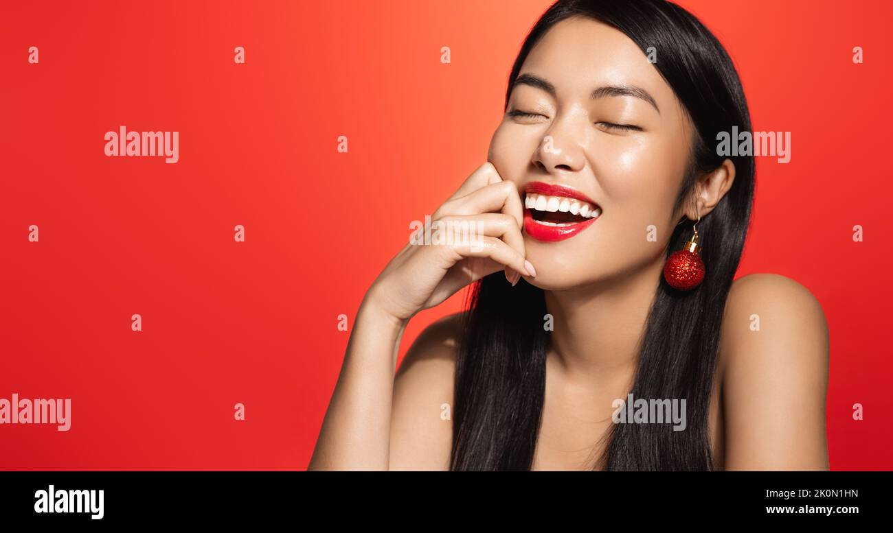 Concetto di bellezza delle donne e delle vacanze. Bella donna asiatica sorridente con gli orecchini di natale di nuovo anno, guardando da parte, toccando la pelle pulita del viso, nuovo anno Foto Stock