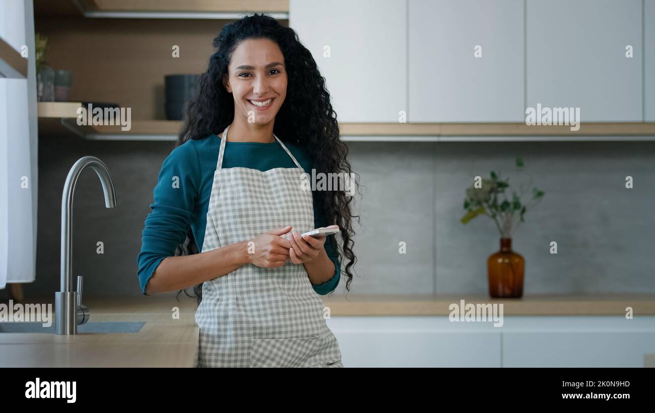 Felice casalinga sorridente arabian hispanic donna madre proprietario di casa con capelli ricci lunghi porta grembiule stand in cucina chat sms fare uso di note di cucina Foto Stock