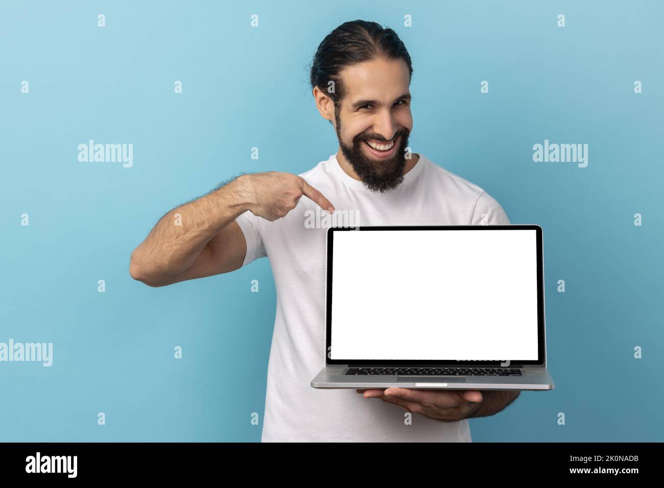 Ritratto dell'uomo con barba che indossa una T-shirt bianca che tiene in piedi il laptop con schermo vuoto e sorridente alla macchina fotografica, pubblicità su internet. Studio in interni isolato su sfondo blu. Foto Stock