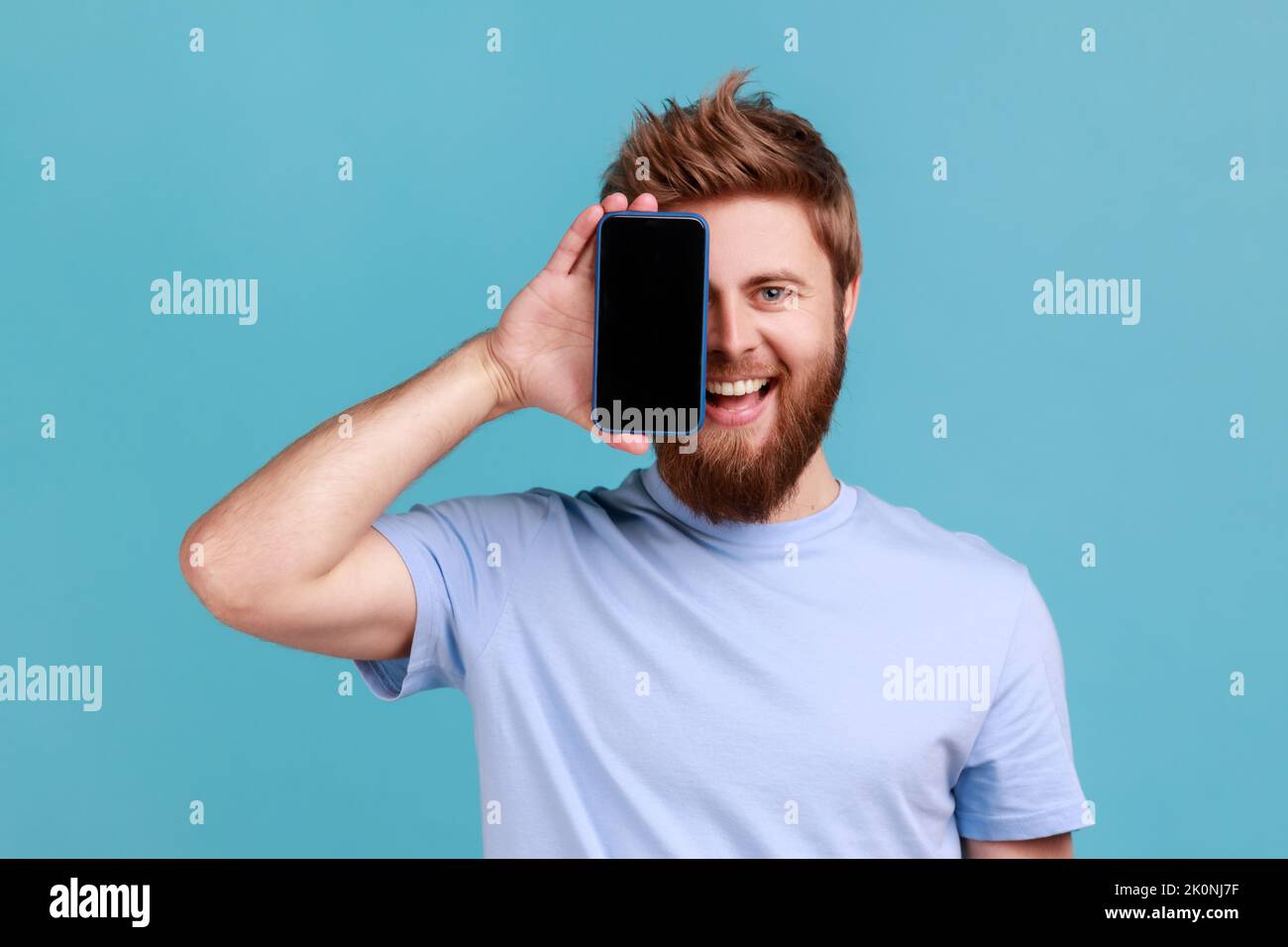 Ritratto di sorridente ottimista uomo bearded coprendo metà faccia con il cellulare e guardando la macchina fotografica con un sorriso toothy, positivo felice faccia. Studio in interni isolato su sfondo blu. Foto Stock