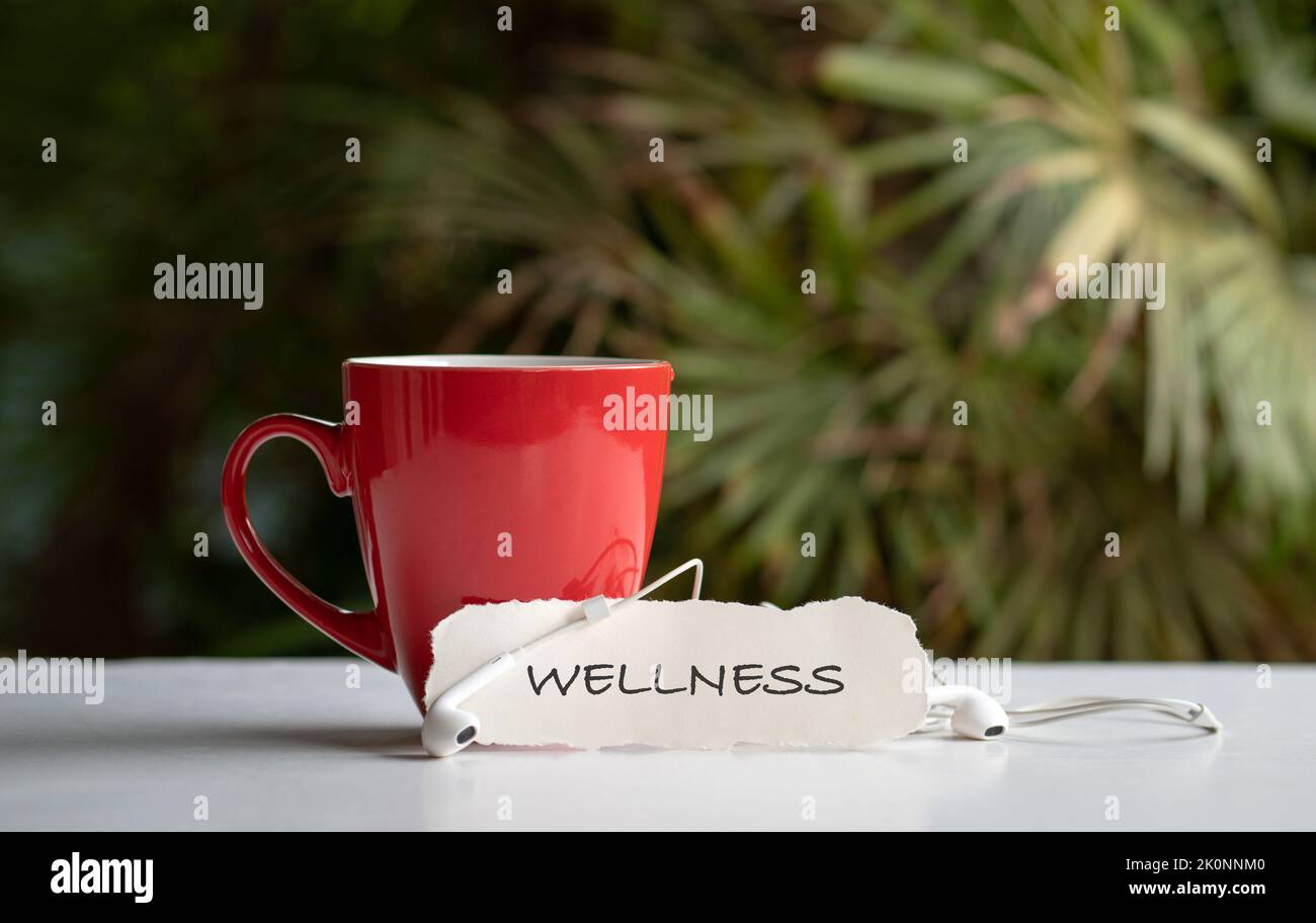 Parola, benessere su un pezzo di carta bianca accanto a una tazza rossa con auricolari. Concetto di auto-cura e benessere. Foto Stock