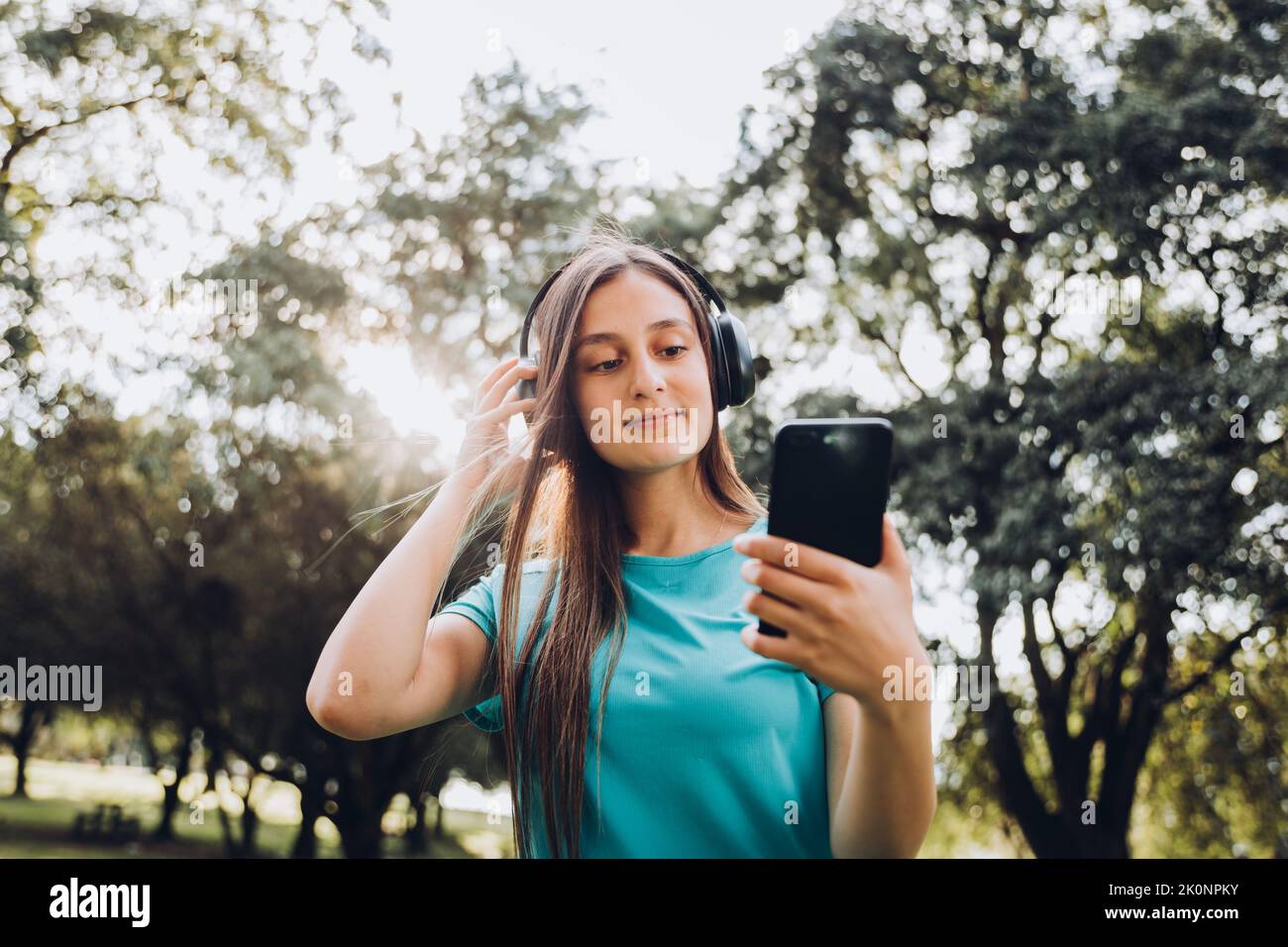 Ragazza adolescente che indossa una t-shirt turchese, con le cuffie, creando playlist sul suo smartphone nel parco. Retroilluminazione del sole Foto Stock