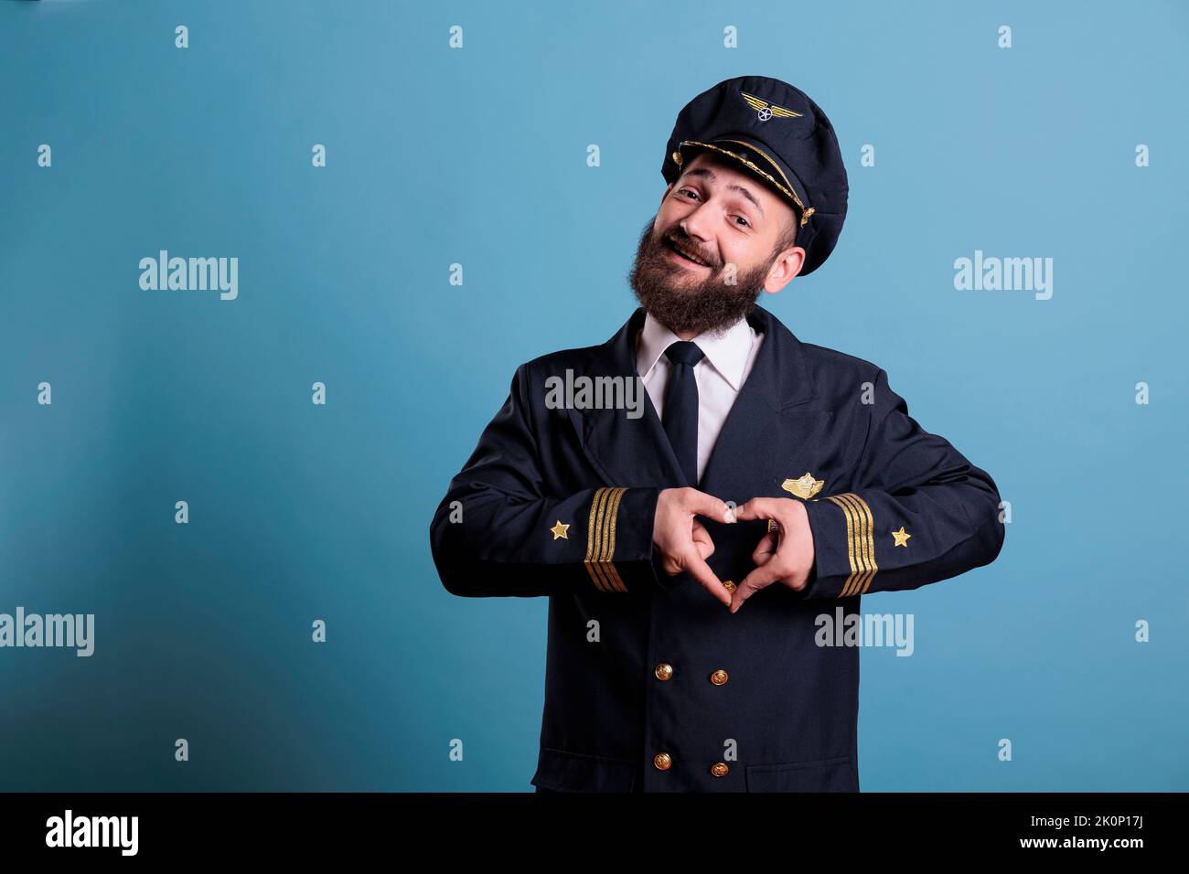 Pilota sorridente di aeroplano che mostra simbolo d'amore a forma di cuore con le dita, gesto romantico concettuale. Capitano amichevole dell'aereo in uniforme esprimente le sensibilità di affetto vista anteriore colpo medio Foto Stock