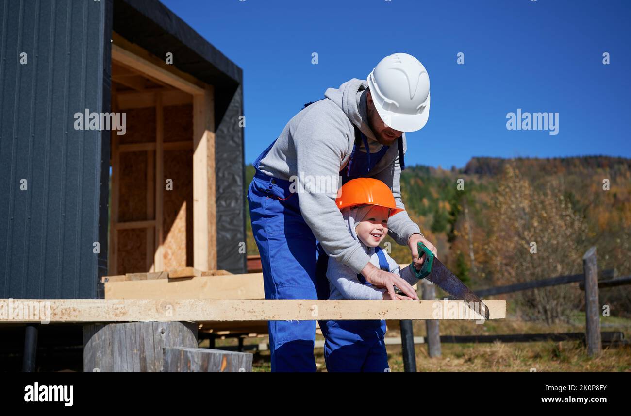 Padre con bambino figlio costruzione legno cornice casa. Costruttori maschi che utilizzano la sega a mano per tagliare tavole in cantiere, indossando casco e tute blu in giornata di sole. Carpenteria e concetto di famiglia. Foto Stock