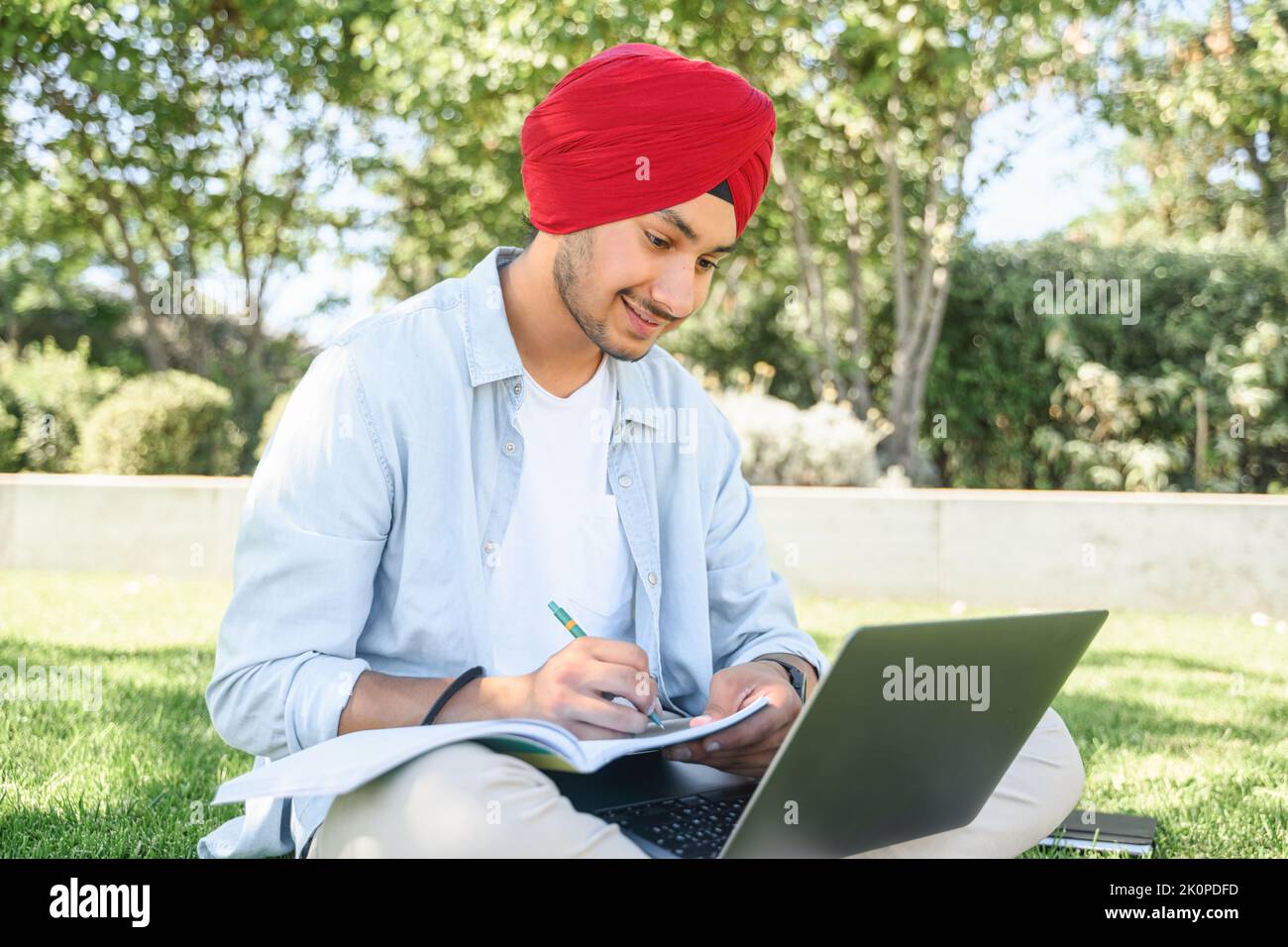 E-learning. Uomo indiano multirazziale in turbante utilizzando il computer portatile per guardare corsi online, webinar, prende appunti seduti nel parco del campus, studente maschile che prepara esami, sostenere corsi di formazione Foto Stock