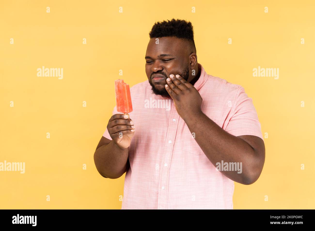 Ritratto di giovane adulto insano che indossa una camicia rosa che tiene un gelato freddo e soffre di dolore terribile, ha denti sensibili. Studio al coperto isolato su sfondo giallo. Foto Stock