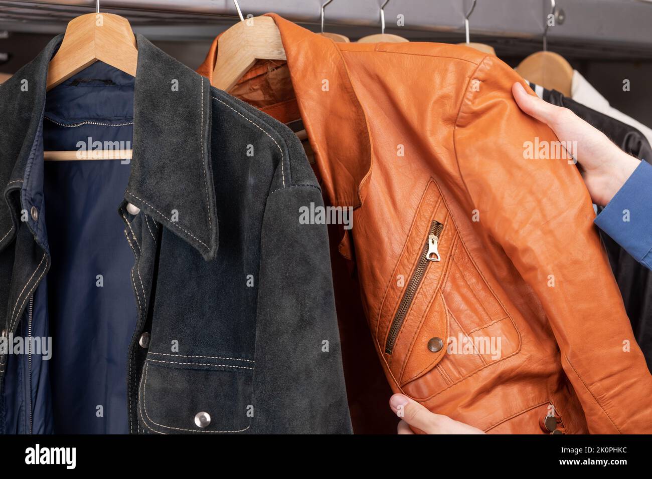 Uomo controlla giacca in pelle arancione in un negozio di seconda mano. Varie giacche d'epoca appendono su una rastrelliera di abbigliamento. Thrifting e sostenibilità in abbigliamento conce Foto Stock