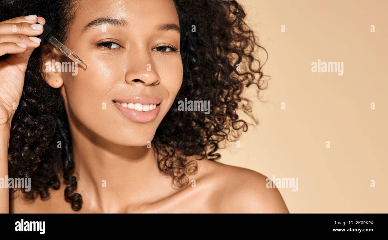 Donna riccia afroamericana che applica olio essenziale sul viso per una pelle idratata utilizzando un contagocce per pipette. cura della pelle del viso con olio essenziale per il trattamento Foto Stock