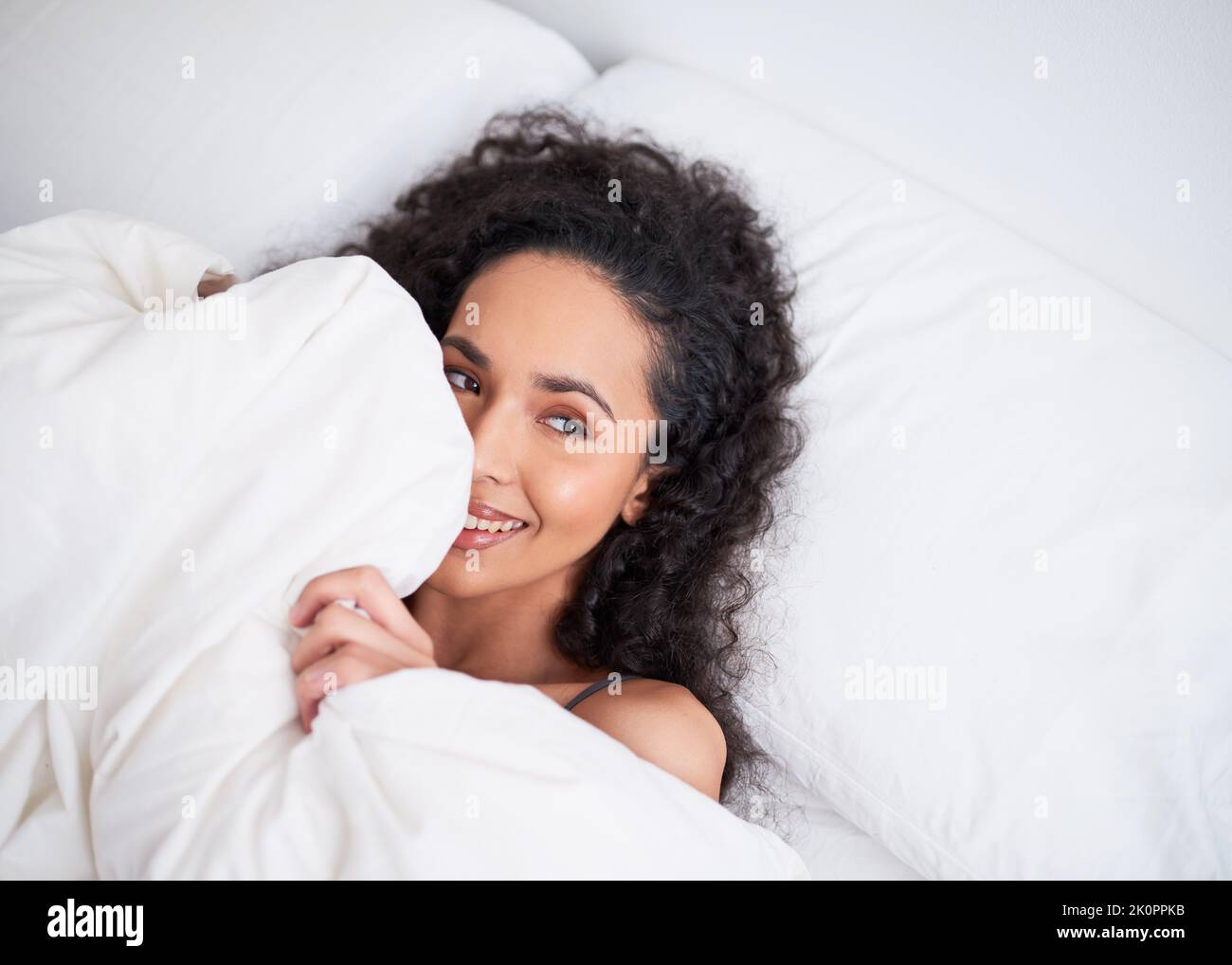 Una giovane donna multietnica si nasconde giocosamente sotto il piumino in lenzuola bianche Foto Stock
