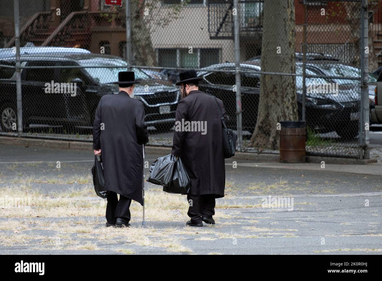 Due ebrei hassidici, possibilmente la famiglia, ritornano a casa dallo shopping prendendo una scorciatoia attraverso un cortile scolastico. A Williamsburg, Brooklyn, New York. Foto Stock