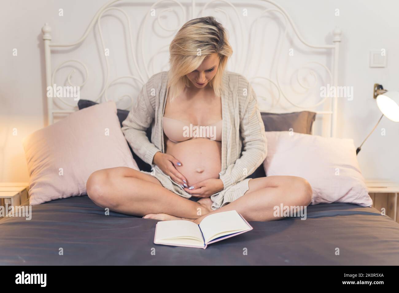 Giovane donna incinta con i capelli biondi seduti su un letto e toccando il ventre mentre legge un libro prima di andare a dormire. Cose da fare quando si aspetta un bambino. Foto di alta qualità Foto Stock