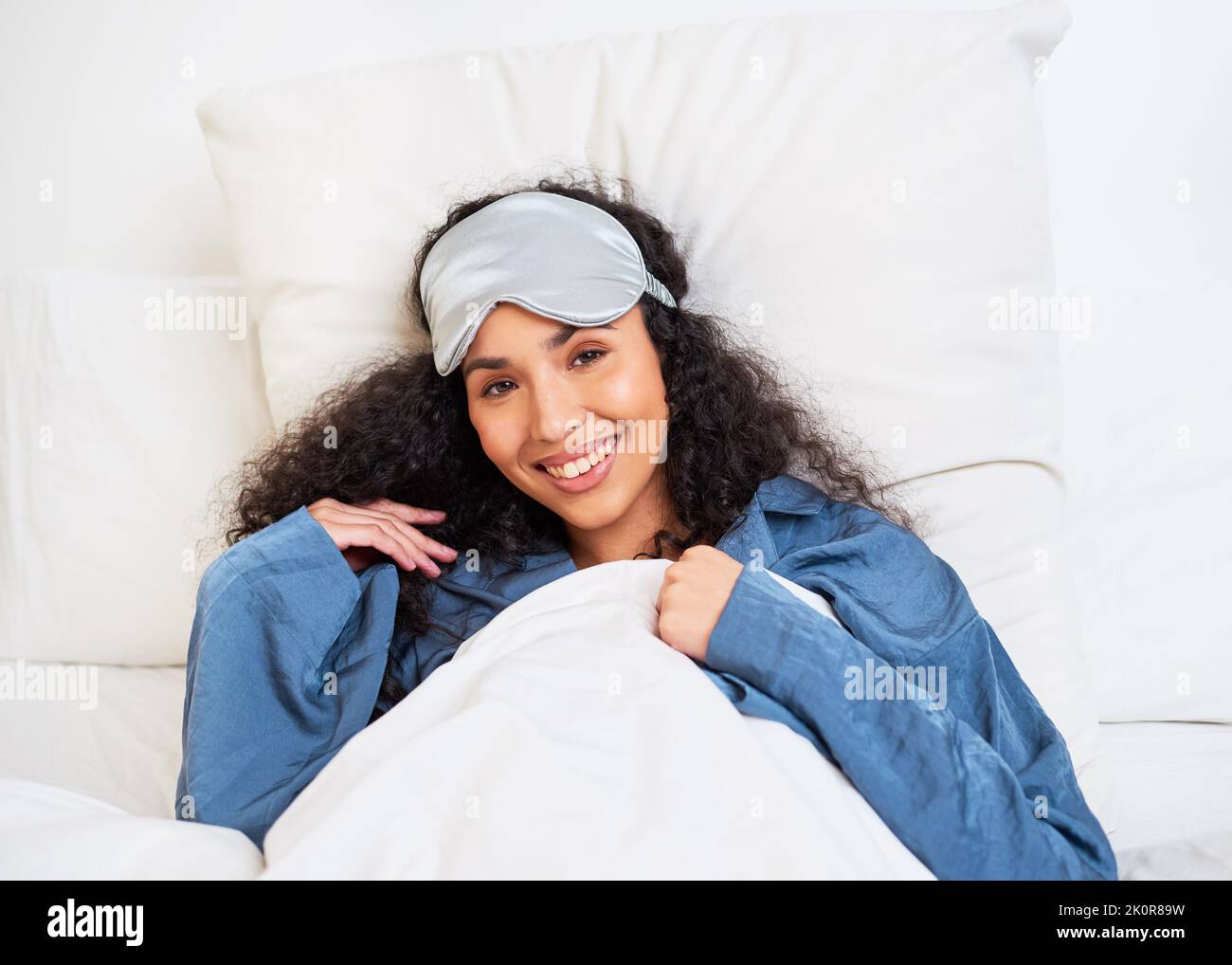 Una giovane donna multietnica si allunga con le braccia dietro la testa e la maschera del sonno Foto Stock
