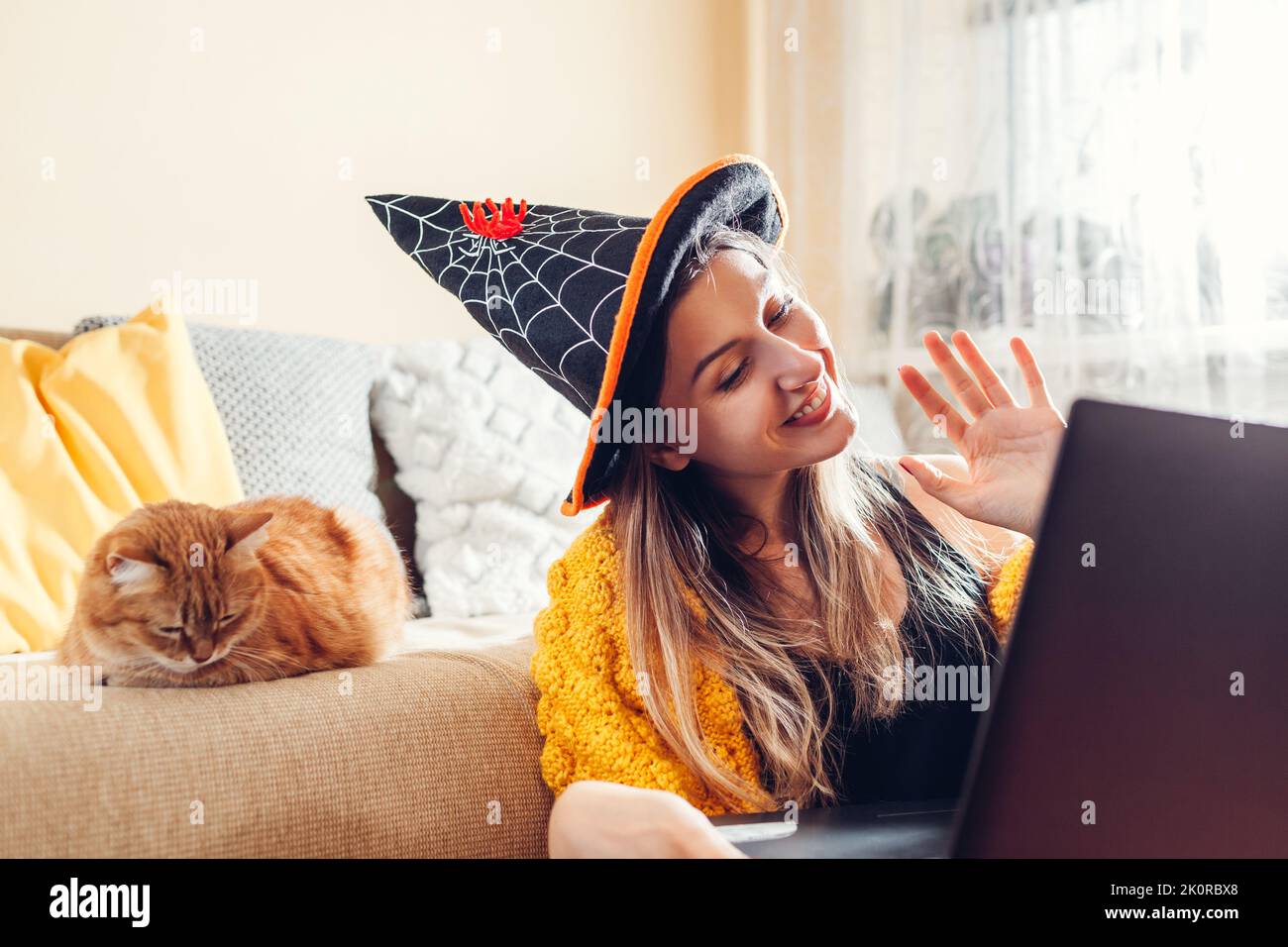 Festa di Halloween a casa. Felice donna sorridente in cappello di strega che svela il hi in linea con la video chiacchierata sul laptop dal suo gatto che dorme nel divano. Restrictio Foto Stock