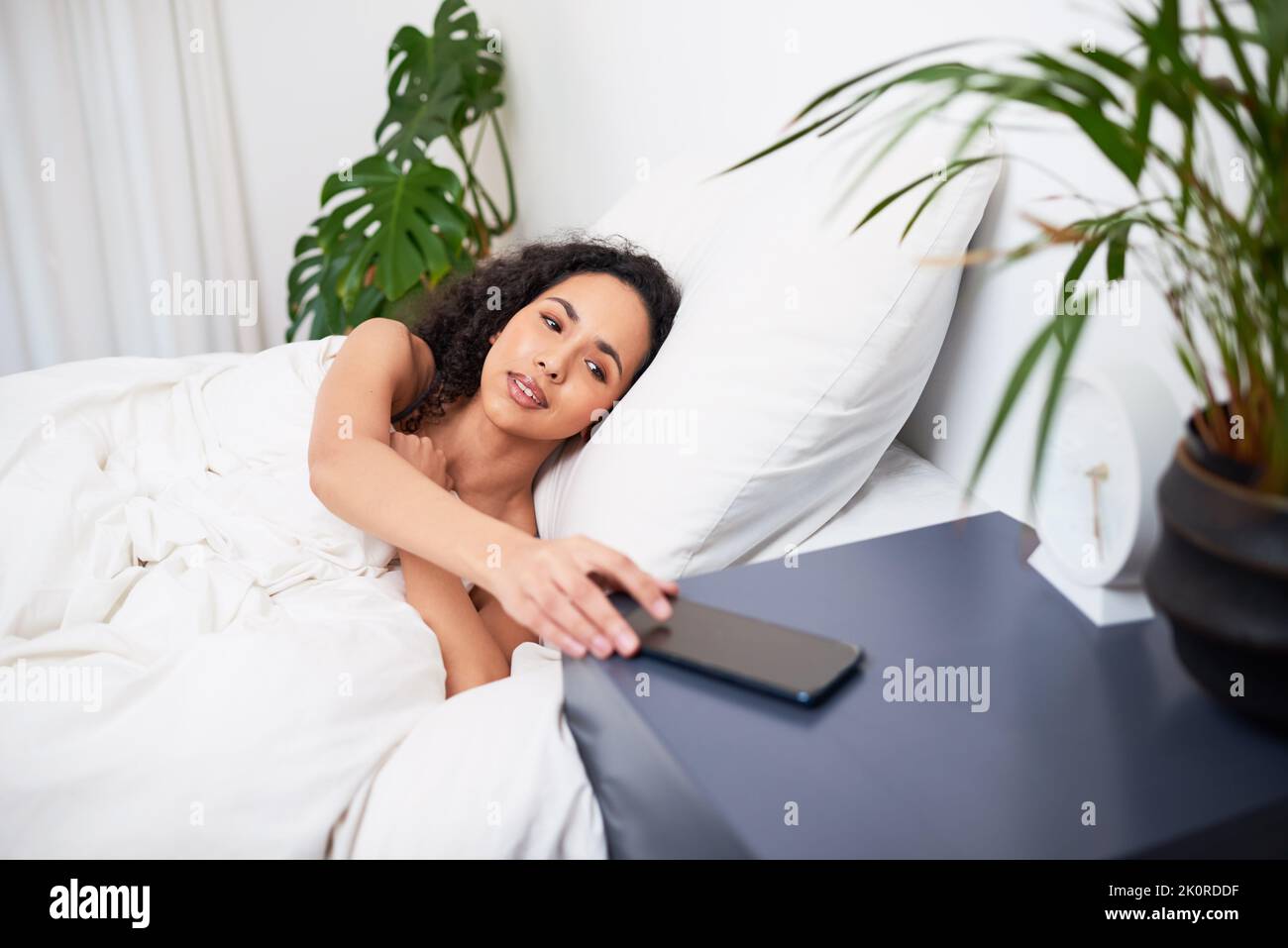 Una giovane donna multietnica si rivolge al cellulare quando si sveglia a letto Foto Stock