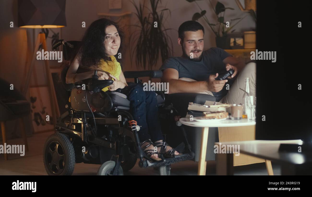 Uomo che gioca in console di videogiochi in TV utilizzando gamepad e donna con una disabilità fisica seduta su una sedia a rotelle a guardare Foto Stock