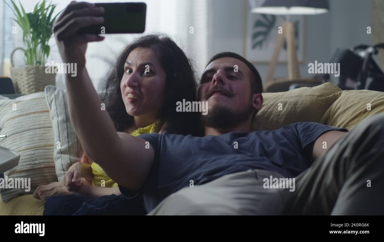 Donna con disabilità fisica e uomo che ride e porta un selfie insieme sullo smartphone per i social network seduti sul divano di casa Foto Stock