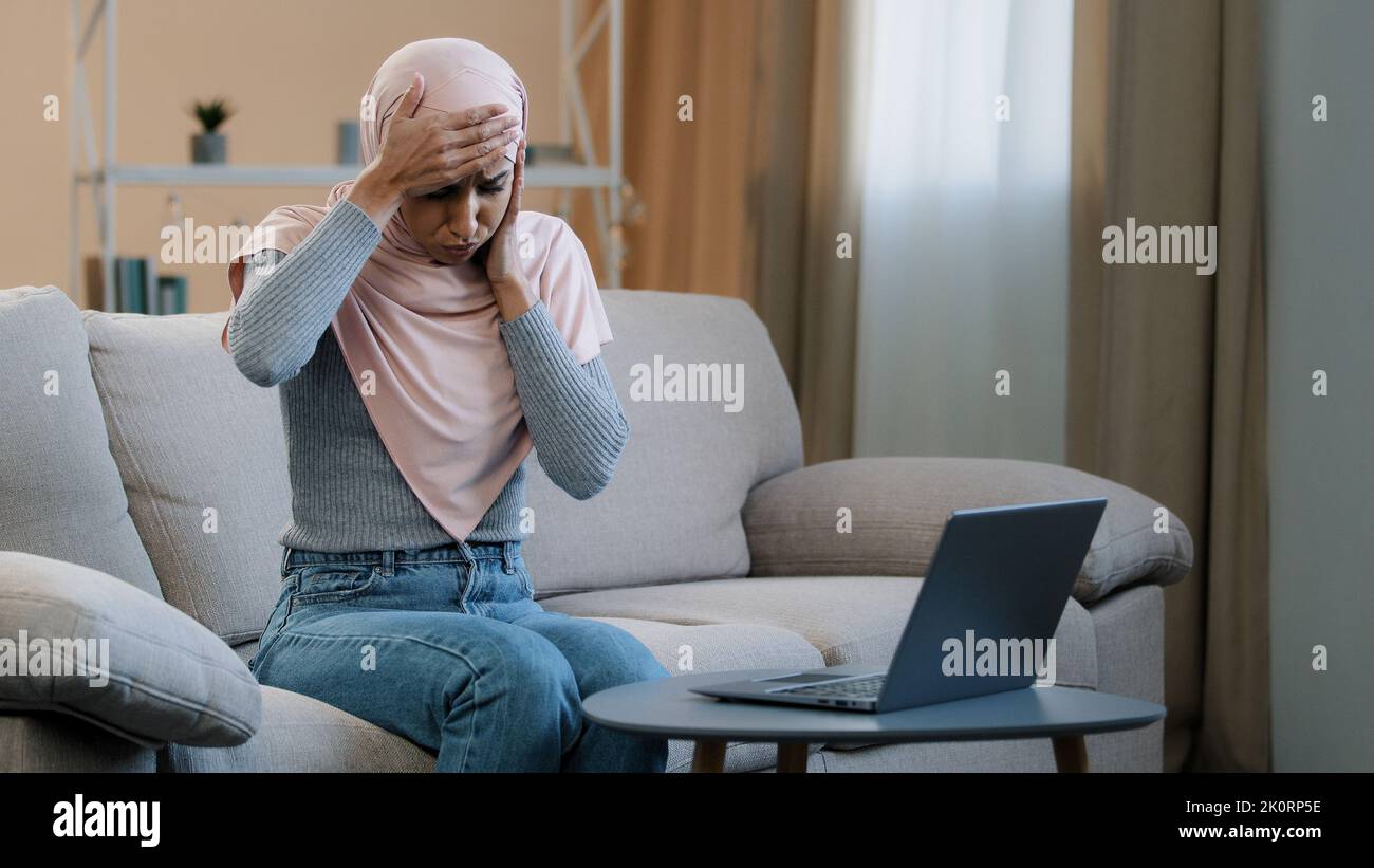Musulmana araba islamica in hijab rosa seduta sul divano di lavoro utilizzando laptop sentire mal di testa stress emicrania grave dolore stanco trista femminile freelancer Foto Stock