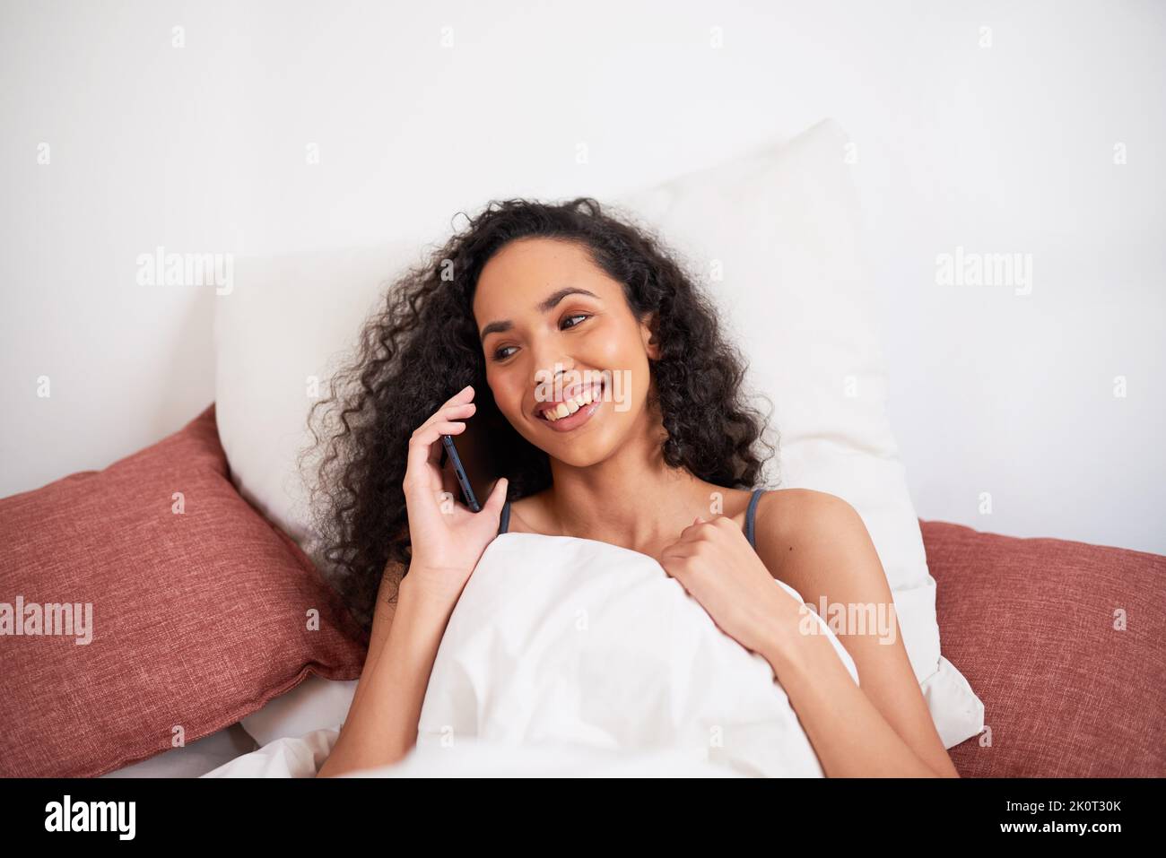 Primo piano di una giovane donna multietnica che chiacchiera sul cellulare in letto con cuscini Foto Stock