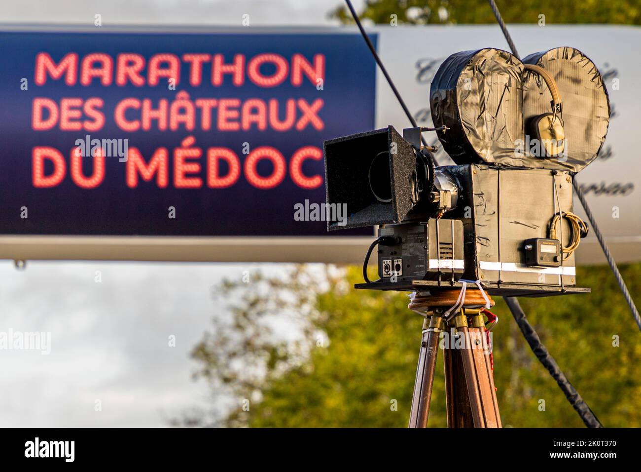 Una fotocamera in cartapesta fatta in casa è posizionata su un treppiede davanti all'insegna illuminata "Marathon des Châteaux du Médoc" a Pauillac, in Francia – un accessorio di costume che corrisponde al tema "Film" del 36° Marathon des Châteaux du Médoc nel 2022. Dalle casse sonore suonano famose colonne sonore cinematografiche. Foto Stock