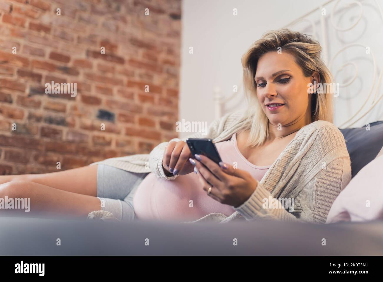 Giovane donna sposata incinta che si rilassa su un letto di notte e lo shopping online sul suo smartphone. Foto di alta qualità Foto Stock