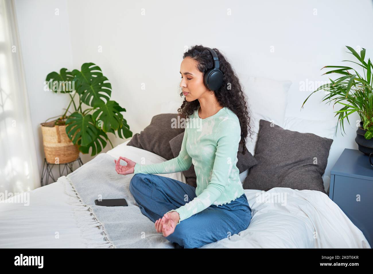 Una giovane donna multietnica siede sul suo letto meditando con l'app di consapevolezza Foto Stock