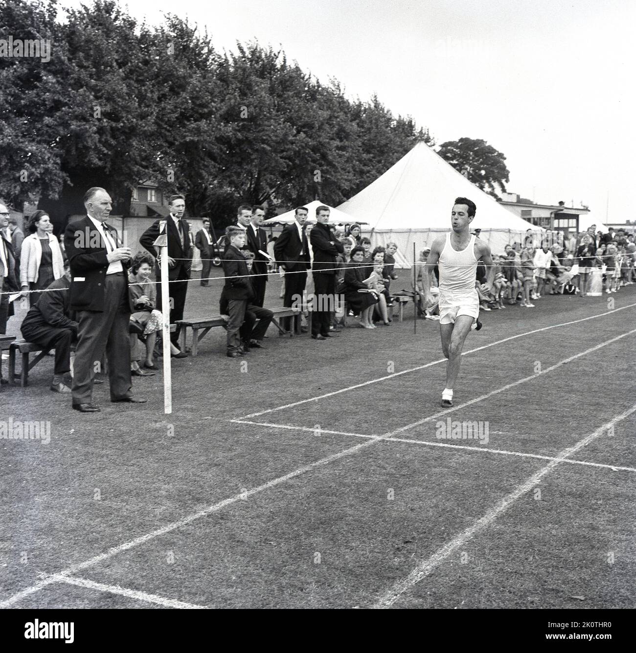 1964, storico, all'aperto su una pista in erba, un atleta maschile con pantaloncini bianchi e giubbotto che gareggia in una gara di corsa, guardato dagli spettatori, Inghilterra, Regno Unito. Foto Stock