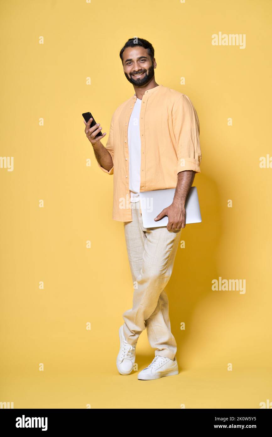 Uomo indiano isolato su un computer portatile di colore giallo utilizzando un telefono cellulare. Verticale a lunghezza intera Foto Stock
