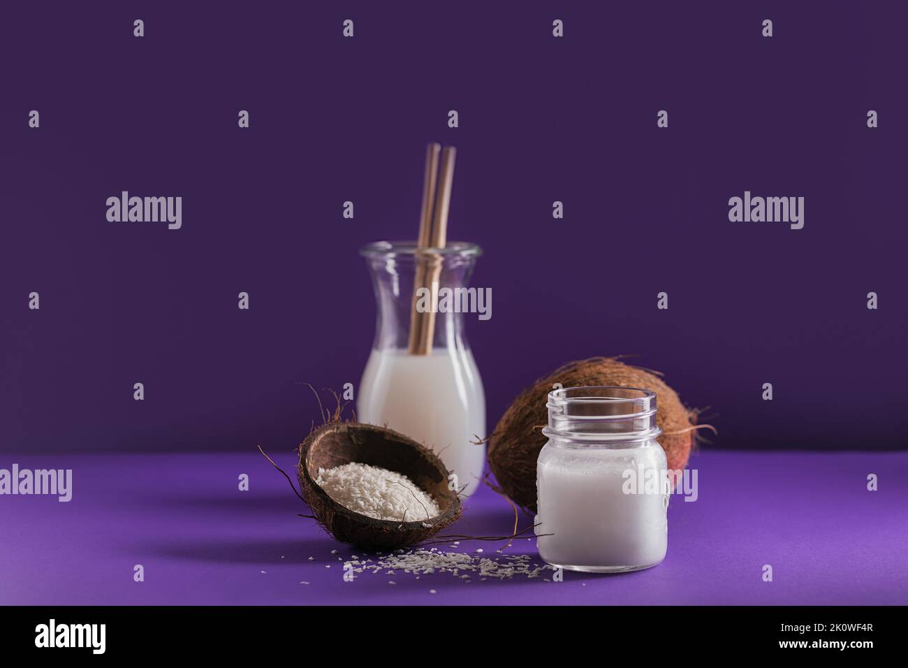 Bottiglia di latte di cocco vegano, olio di cocco, cocco intero e fiocchi su sfondo viola Foto Stock